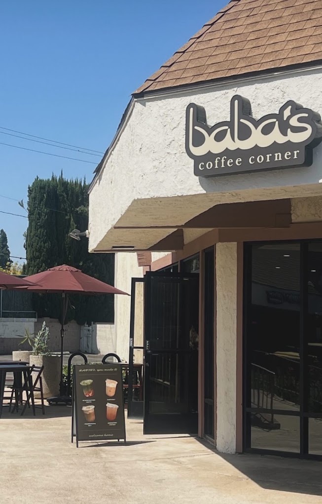 Baba's storefront from the patio — the sign, a red umbrella over bar tables, a young olive tree and the A-frame menu board.