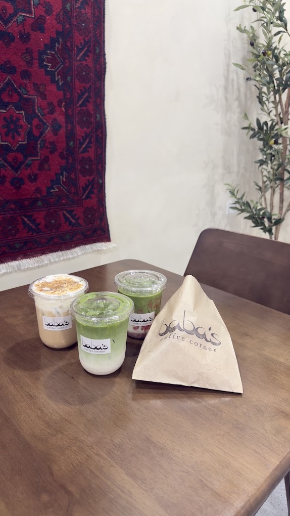 Three Baba's drinks and a kraft pyramid bag on a wood table, a deep-red Afghan rug hung on the wall and an olive tree behind.