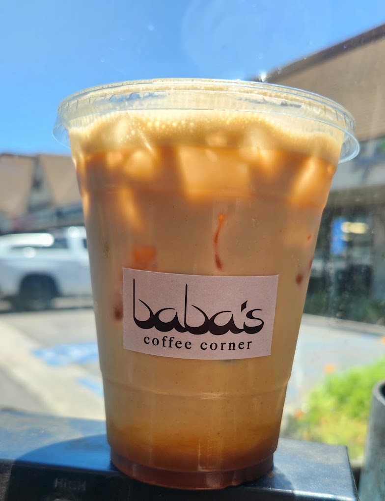 A hand holding an iced Baba's Latte, its kraft label facing out, with the plaza and blue sky behind.