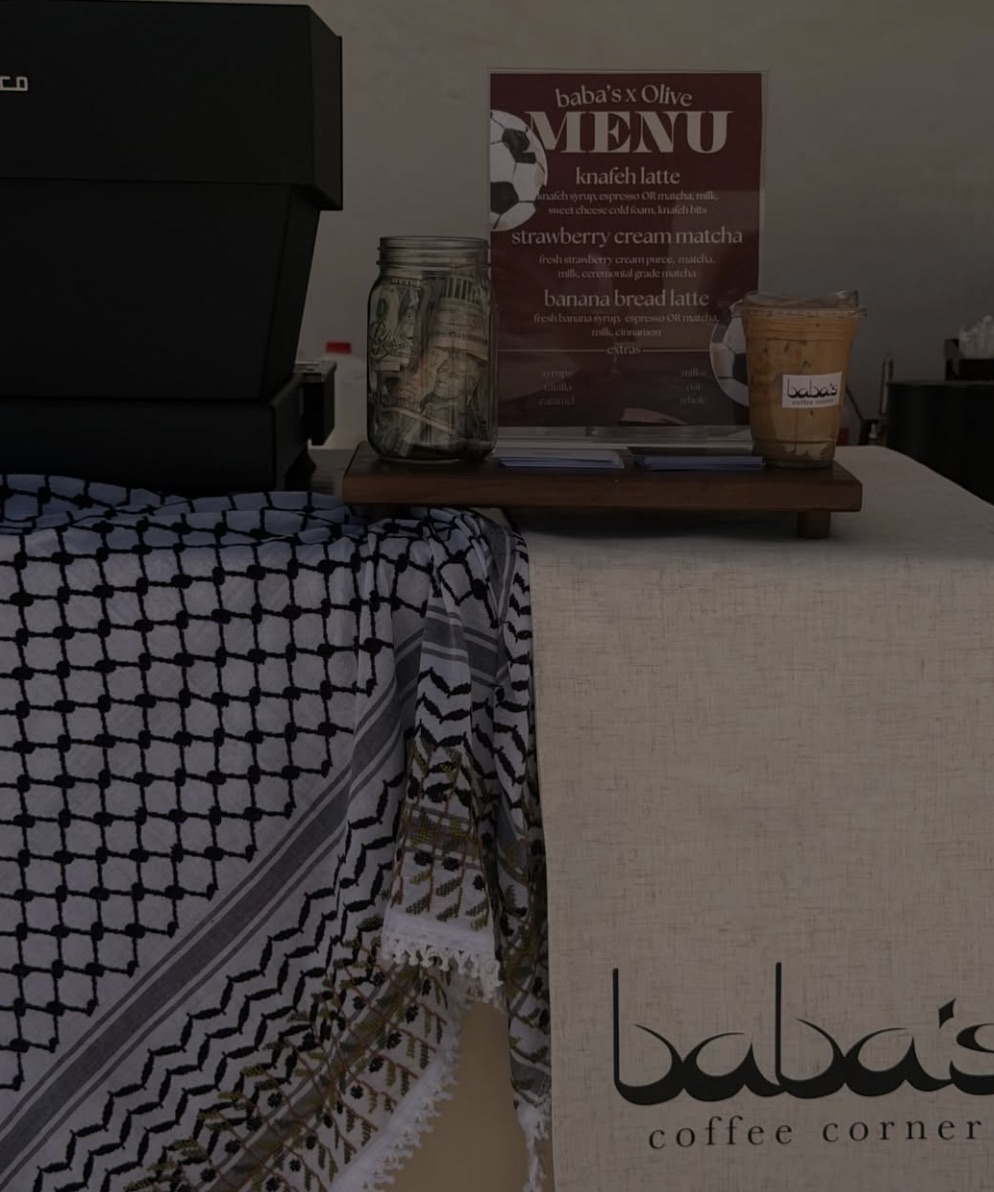 A black-and-white keffiyeh folded over the counter beside the oxblood 'baba's x Olive' menu board and a mason-jar tip jar, next to the espresso machine.