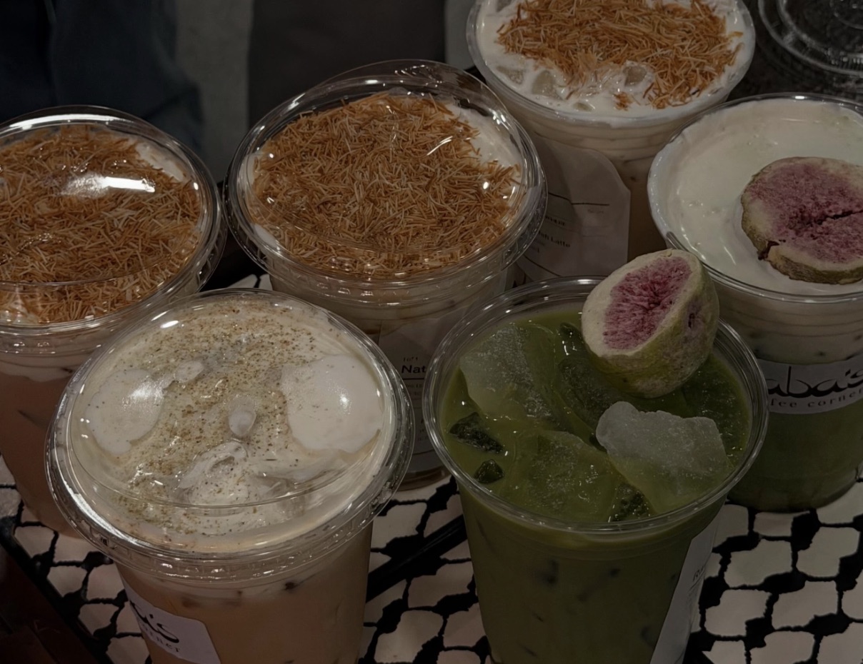 A cluster of Baba's iced drinks topped with toasted knafeh crunch and a fig-garnished matcha, set on a black-and-white keffiyeh-patterned surface.