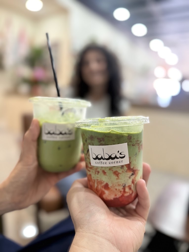 Two hands raising Baba's iced matcha drinks together, warm bokeh behind.
