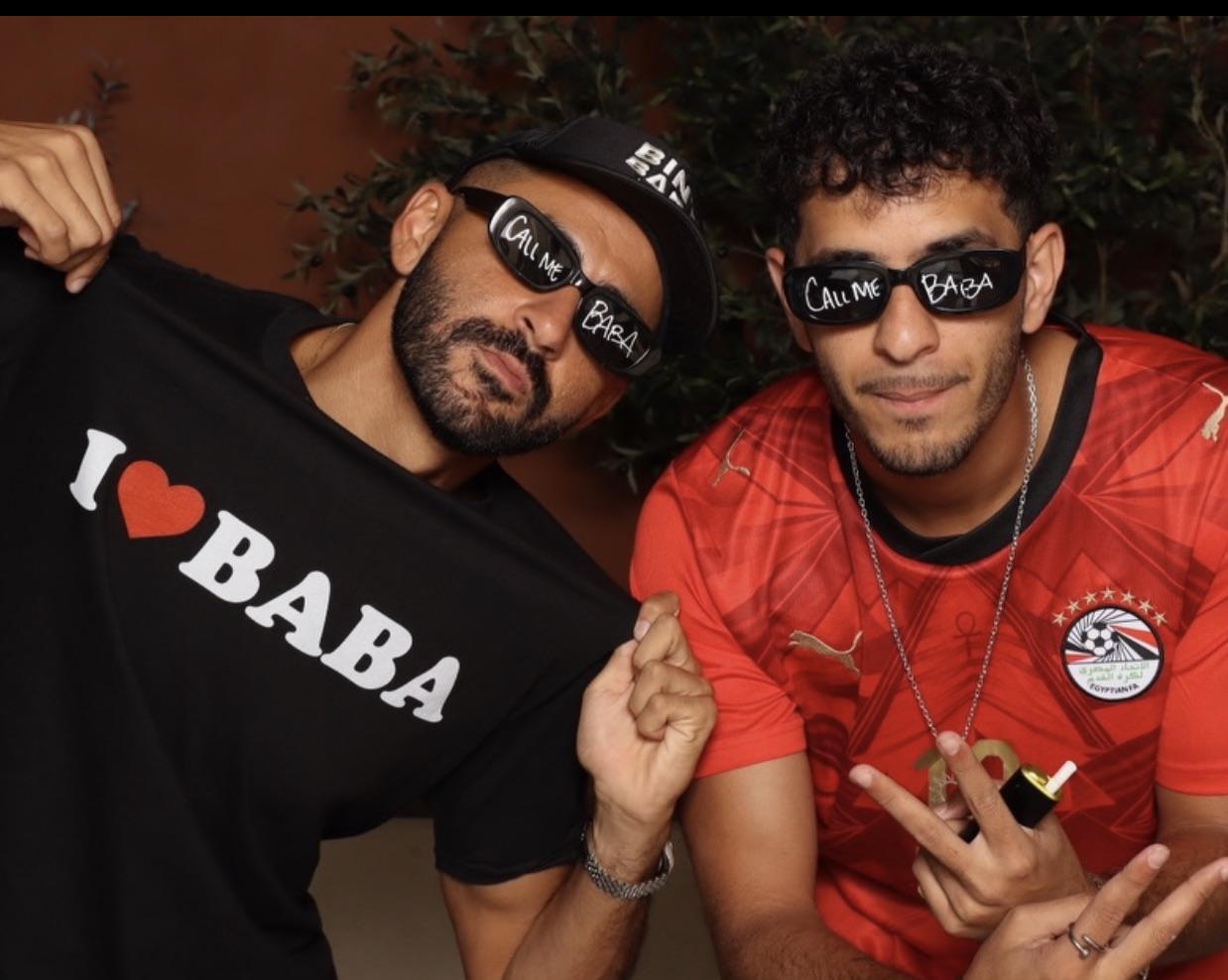 Two of the family holding an 'I ♥ BABA' tee, wearing 'Call Me Baba' sunglasses, against a terracotta wall with olive branches overhead.