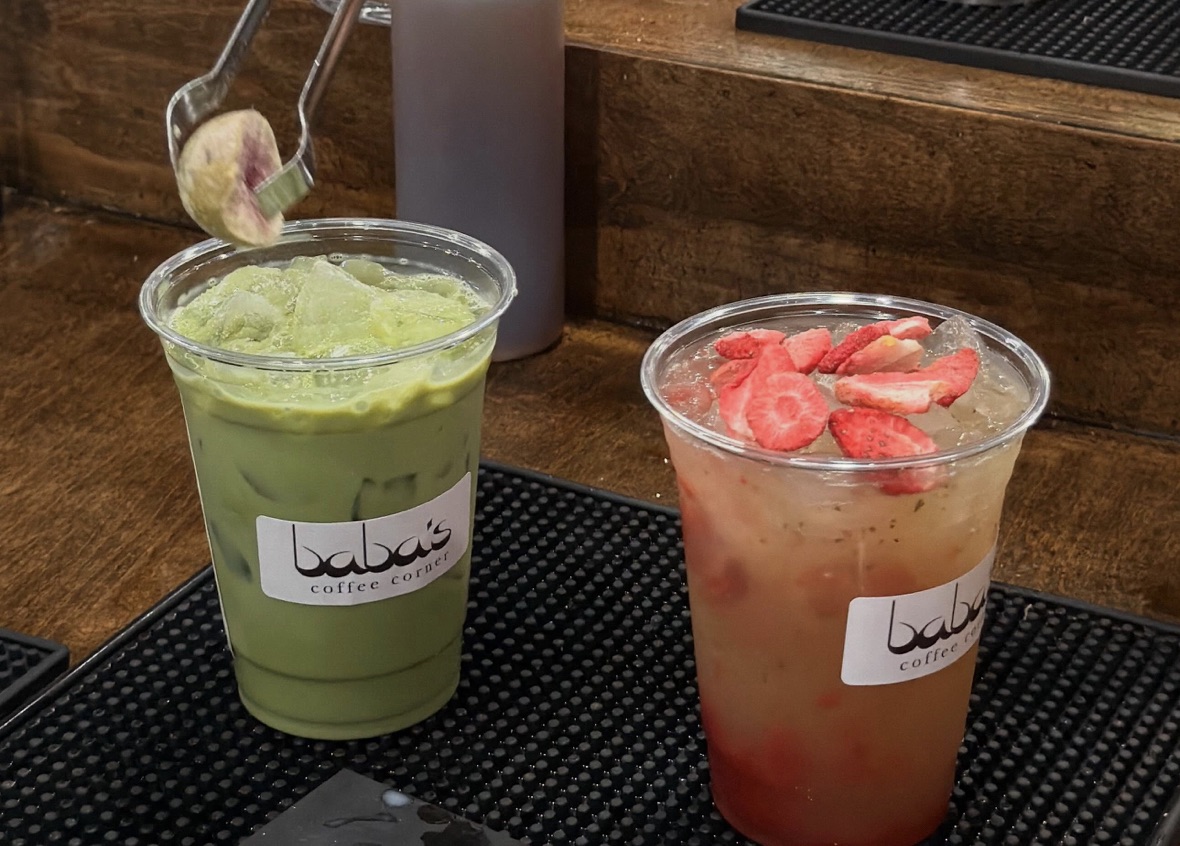 An iced matcha and a strawberry refresher side by side on a black bar mat against a warm wood counter, a fig on a spoon behind.